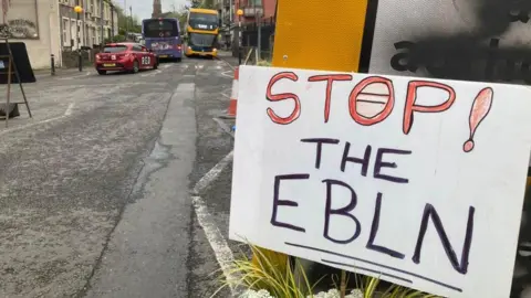 A sign which says "STOP! THE EBLN" positioned a graffitied road sign on a main road in Barton Hill. Two buses and a red car can be seen driving down the road.