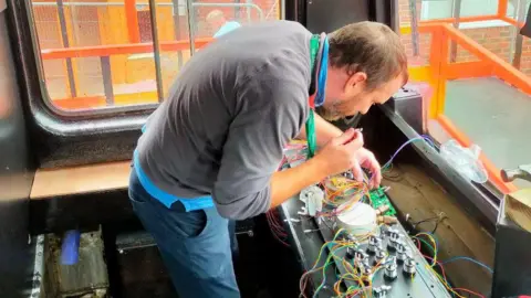 The Dales School An engineer, who wears a grey jumper, blue top and jeans, works upon a switchboard in the driver's cab of a Pacer train. The train is located in the playground of a school yard.