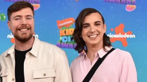 Getty Images MrBeast pictured with Ava Kris Tyson at the Nickelodeon Kids Choice Awards. MrBeast has short brown hair and a short beard and wears a white overshirt over black T-shirt. Ava has shoulder-length brown hair parted to the side and wears a pink shirt. 