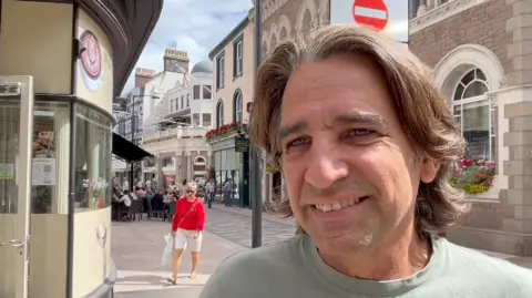 Ian Carr is standing in the foreground of a pedestrianised town or city street, with shops, cafés and historic-looking stone buildings visible behind.