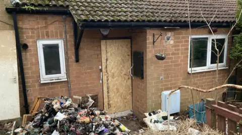 A red brick bungalow which has been damaged by fire - the front door is boarded up and there is a large pile of debris beneath the window on the left of the image