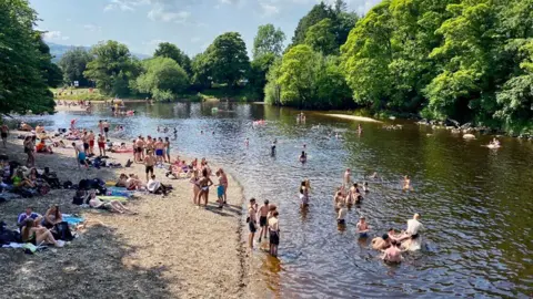 Yorkshire Water A section of the River Wharfe in Ilkley, looking down from the bridge over New Brook Street. It is a summer day and there are lots of people sat on the river bank and swimming in the water. 