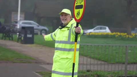 Ian Preston pictured smiling holding a yellow and red stop sign. He wears an illuminous yellow jacket.