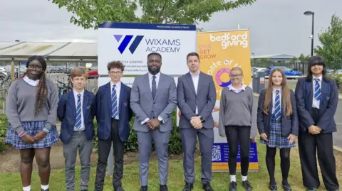 Bedford Giving Six pupils standing outside their school, with two adults in the middle. All the pupils are wearing grey and blue school uniform, and the men in the middle, grey suits.