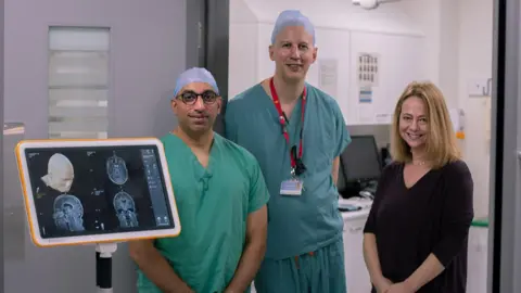 PA Media Three people standing in a line in a hospital room. Two men are in teal scrubs with blue hairnets on their heards. One is short with glasses, the other taller. The third person is a woman wearing a loose black dress with shoulder-length blonde hair. All of them are smiling at the camera.
