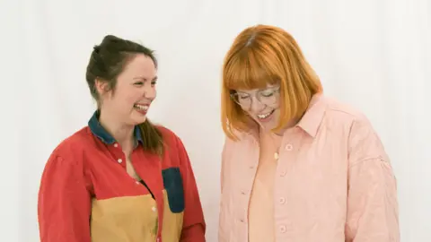 Kerry Curl A colour mid-length photo of two women pictured from the waist up, standing against a plain white backdrop. On the left is a woman in her mid-30s, with long, dark brown hair tied back and then swept forward over her left shoulder. She is wearing a long-sleeved top with red shoulders and sleeves and a mustard-coloured middle section, with a dark blue pocket patch over the left side of her chest. She is looking and smiling, laughing, at the woman standing next to her. She is also in her mid-30s, with shoulder-length red hair worn loose, wearing glasses, and a pale pink top over a pale peach-coloured t-shirt beneath. She is also smiling but looking down at the floor.