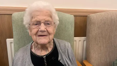 BBC Hazel, who is woman with white hair and glasses, is looking at the camera with a slight smile on her lips. She is wearing a black top and silver sparkly cardigan. She is sitting on a green chair and there is a brown empty chair next to her. 