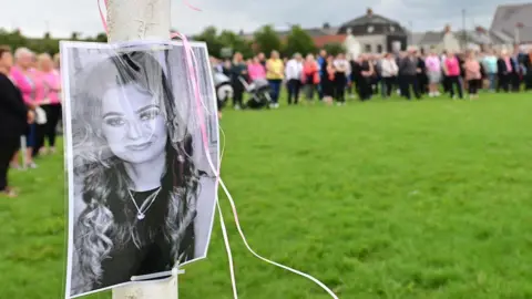 Pacemaker Press A black and white image of Chloe Mitchell tied to a lamppost in a park. There is a group of people standing in the grass.