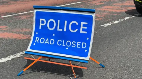 A blue sign with white writing. The writing says 'Police Road Closed'. The sign sits on orange legs on a road which has white and red markings