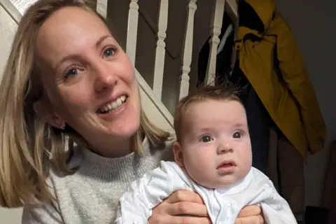 Louise smiles at the camera, holding her baby