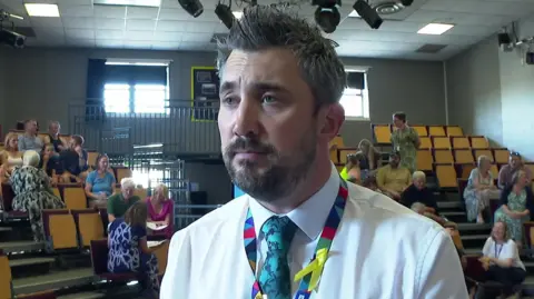 Principal Gareth Smith wearing a white shirt, a patterned tie and a colourful lanyard with a yellow ribbon attached. He is standing in an indoor auditorium with tiered seating and several people sitting in the background. 