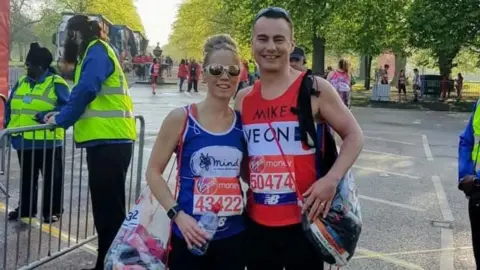 Jo Wood Jo and Mike wearing charity T-shirts, with race numbers pinned to them, at the red start of the London Marathon. They are each carrying a see-through bag with kit inside. There is a line of green trees in the background.