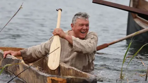 Operation Phoenix A laughing man wearing a beige tunic, waist-deep in water, towing a log boat and holding a wooden paddle