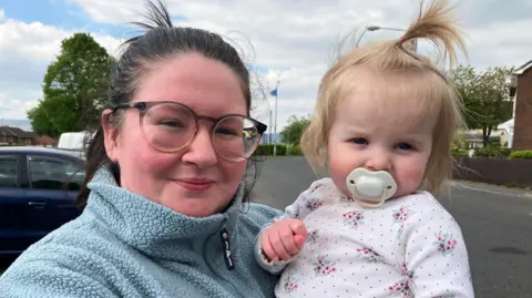 A woman with black, pulled-back hair is standing on a street, holding a small child in her arms on a street. She is wearing black-rimmed glasses and a light blue fleece. The young girl is wearing a white floral top and has a white dummy in her mouth. Her blonde hair is also tied up.