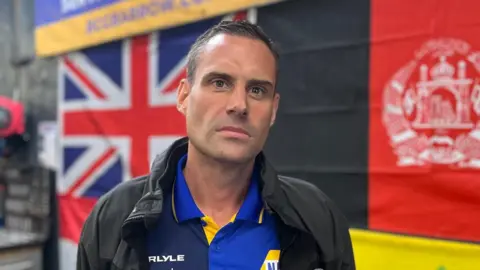 Thomas Henderson is clean shaven with short, dark hair and is wearing a black anorak-style jacket and polo shirt in navy, royal blue and yellow. He is standing in front of a wall of different nationalities' flags.
