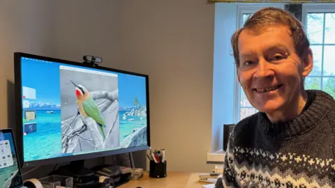 BBC/Ed Hanson Stan Jenkins, on the right hand side, wearing a grey patterned jumper smiling. He has grey hair and sitting at a computer desk. On the left hand side is a computer monitor with a green and red bird with a long black beak picture on it. There is a pen pot on the table. 