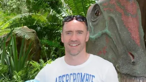 BBC Paul Doyle wearing a white t-shirt, smiling with sunglasses on his head.
