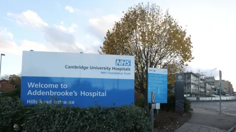 Exterior image of Addenbrooke's Hospital. In the foreground is a sign welcoming people to the hospital's Hills Road entrance. In the background are hospital buildings.