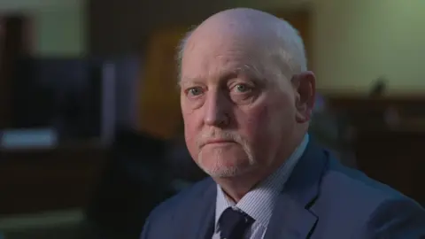 A man with a short grey goatee beard and balding grey hair looks sideways towards the camera. He is wearing a navy suit, striped shirt and navy tie. The background is blurred, but shows an old court room. 