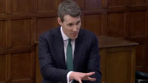 House of Commons A man with brown hair in a suit gestures towards MPs who had brought complaints about Cil to a debate in the wood panelled committee room in Westminster Hall.