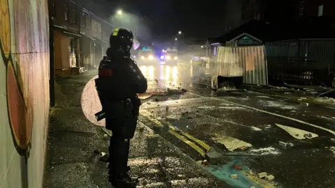 BBC Police officer in riot gear standing looking at a street covered in debris, the lights of police landrovers in the background