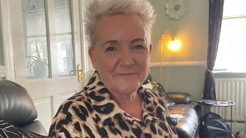 Lorraine Beavers MP with short blond hair and wearing a leopard print top sits smiling on a sofa in a house.