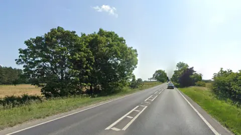 Google The A66 is a single carriageway with white road markings depicting 'no overtaking' down the middle. The road is bordered by some lush green trees and fields beyond. There is a grey car travelling on the right-hand side of the road.