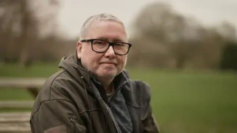 The subject is sitting down, wears glasses and a brownish/green hooded jacket. 