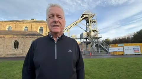 Jim Scott/BBC Ian Lavery looking into the camera and smiling. He is standing in front of Woodhorn Colliery which is a stone building with an old bit of mining equipment with is a large metal machine with a wheel on top outside. He has grey short hair and is wearing a black pullover sports top with a shirt underneath.