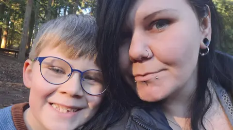 Saxon and his mother Cara. Saxon is wearing blue glasses and is smiling. Cara has black hair covering one eye and has piercings in her nose and lower lip. They are standing in a wooded area.