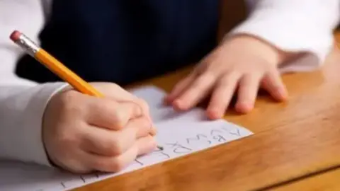 A child draws out letters on a piece of paper against a wooden table. The pencil is yellow and she writes with her right hand.