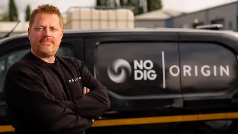 Origin Tech Co-founder of Origin Tech, John Marsden, with red hair, moustache and stubble. He is wearing a black jumper and standing with his arms folded. In the background is a black van with the logo 'No Dig' 'Origin'. 