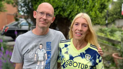 Gary and Jan pose for a photo. Gary has his arm around Jan's shoulder. Jan has long blonde hair, while Gary is bald. They both wear Leeds branded merchanise.