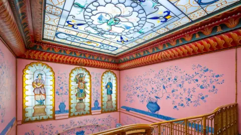 Brighton & Hove Museums A pink room with pretty blue birds and flowers painted on the walls. The ceiling appears like it is made of pale glass. The sun is shining through it. 