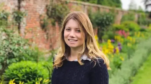 Rebecca Ableman smiling to the camera in a dark navy knitted jumper - there is a beautiful walled garden behind her