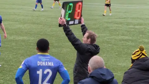 supplied An assistant referee holds up an electric sign on the side of the football pitch signifying a substitution. A player in a blue strip with a number 12 on his short stands waiting to come on.