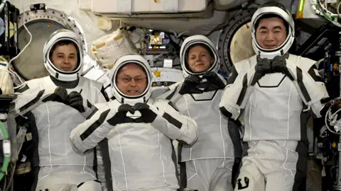 Four astronauts wearing white spacesuits and helmets pose for cameras smiling and making heart shapes with their hands and fingers. They are surrounded by wires and other equipment inside the International Space Station.