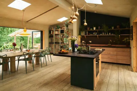 Channel 4 A shot of the interior of the house. A kitchen area which has wooden drawers and black counter top. In the kitchen there is shelving with plant pots, books and kitchenware. A bowl of fruit, flowers and cooker top sit on the counter of the island. Beside there kitchen there is a wooden table with green chairs and a book case.