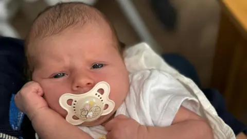 Ami Boyd Flossie Bilboe is pictured as a newborn baby. She is wearing a white baby grown and has a dummy in her mouth. 