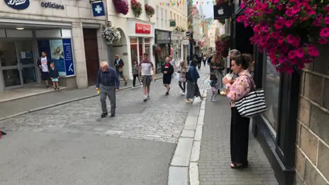 Guernsey's St Peter Port Town High Street. A woman is standing drinking a milkshake under a hanging basket of purple flowers. On the cobbled street, people are walking to and from the camera vantage point. Between the buildings on either side of the cobbled street is multi-coloured bunting. 