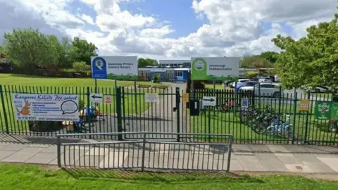 Google Street View Google Street View image of the outside of the school, with signs and metal fences and grass surrounding it.