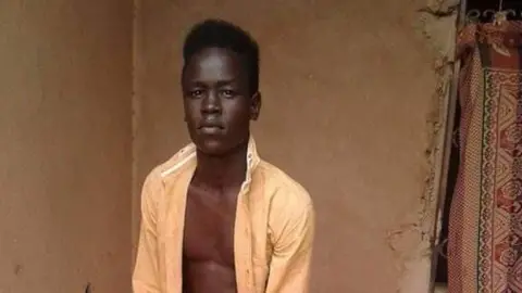 Andrew Kueth A young man with black hair and a beige, unbuttoned shirt in a brown plastered room with a decorative rug on the wall.