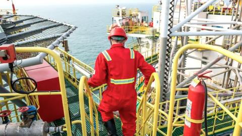 A worker dressed in a red protective coverall with reflective yellow stripes and a red hard hat is seen from behind walking down a yellow metal staircase on an offshore oil platform. The platform is equipped with various pipes, railings, valves, and machinery, extending out over the ocean visible in the background. A red fire extinguisher is mounted on the right side of the image, highlighting safety measures on the industrial site.