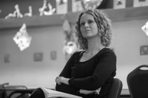 Anna Simpson Anna is sitting down inside, perhaps a studio, wearing a black top, with a pile of papers or folder on her lap. The photo is taken from a lower level, as she looks beyond the camera. She has curly hair and she is pursing her lips slightly like she is concentrating.