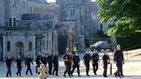 PA Media About 14 police officers wearing dark clothing and police caps walking outside on the grounds of Buckfast Abbey.
