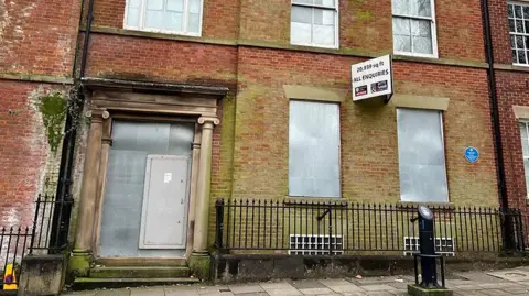 Paul O'Gorman/BBC The Georgian home of prominent suffragette, Edith Rigby and her husband Charles on Winckley Square. It is red brick, stained with green, the  front door has stone pillars either side and is boarded up with metal, as are two windows next to it. There are black railings on the street with basement windows just visible behind