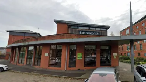 The Bluebrick building is on the left and in the centre, with the sign visible. The building on the right has five storeys. Three cars are parked in the foreground.