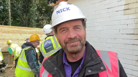 Laura Foster/BBC Presenter of DIY SOS Nick Knowles wearing a white hard hat and pink hi-vis vest