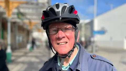 BBC/George Carden A man, with glasses, wearing a cyclist helmet and a blue jacket. He is standing outside a train station.