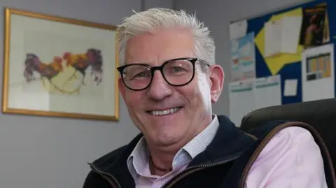Alex Dunlop/BBC Mark Gorton, a man sitting down who is looking directly at the camera and smiling. He is wearing a pink shirt with a gilet over the top.